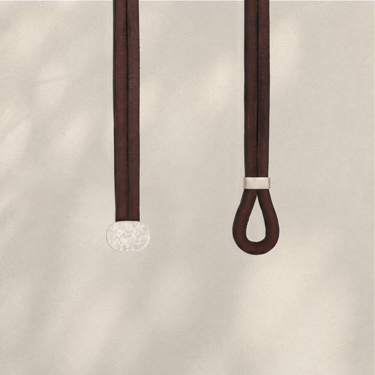 Two dark brown leather strap accessories hanging against a light beige textured background. One strap ends in a round textured silver disc, and the other has a small silver band near the top of a looped end.