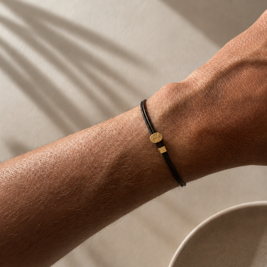 A close-up of a person's wrist wearing a minimalist black cord bracelet with a small 14k solid gold geometric charm, set against a warm beige background with soft leaf shadows.