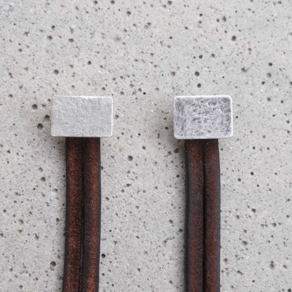 Two rustic brown leather cords with square silver-toned metal end caps, laid flat on a speckled light gray surface. One is aged and has some patina, the other one is new and shiny.