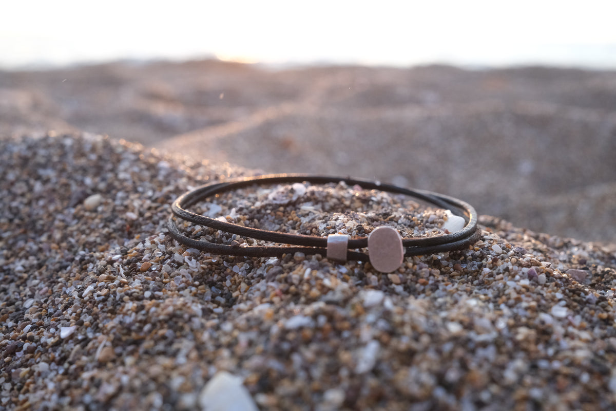 A dark brown leather bracelet with a silver-toned toggle clasp and a small flat metal disc charm, resting on sandy beach gravel with a blurred sunset background.