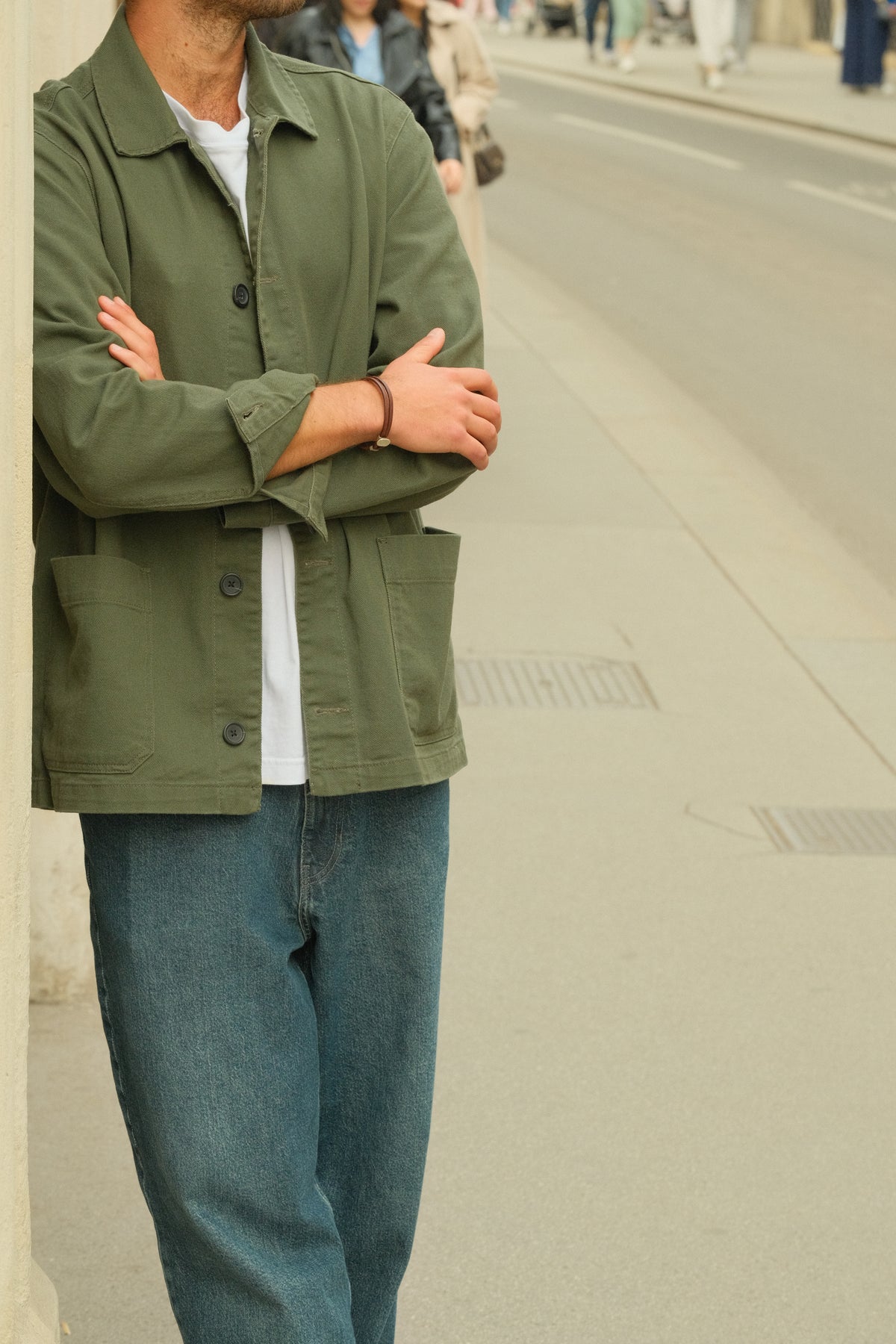A man wearing an olive green utility jacket over a white t-shirt and blue jeans, standing on a city sidewalk with his arms crossed. wearing a silver leather armband.