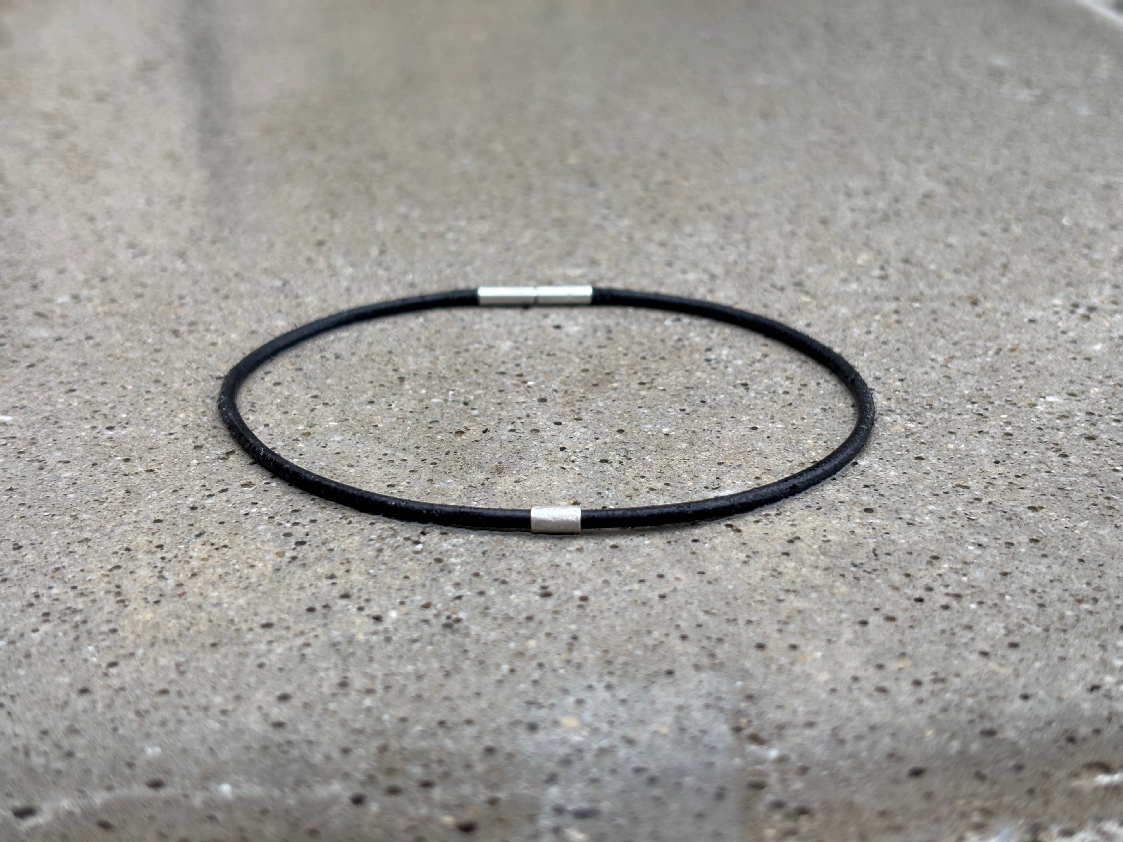 A minimalist black cord anklet with two small silver metal end caps, resting on a textured gray concrete surface.
Baionett clasp