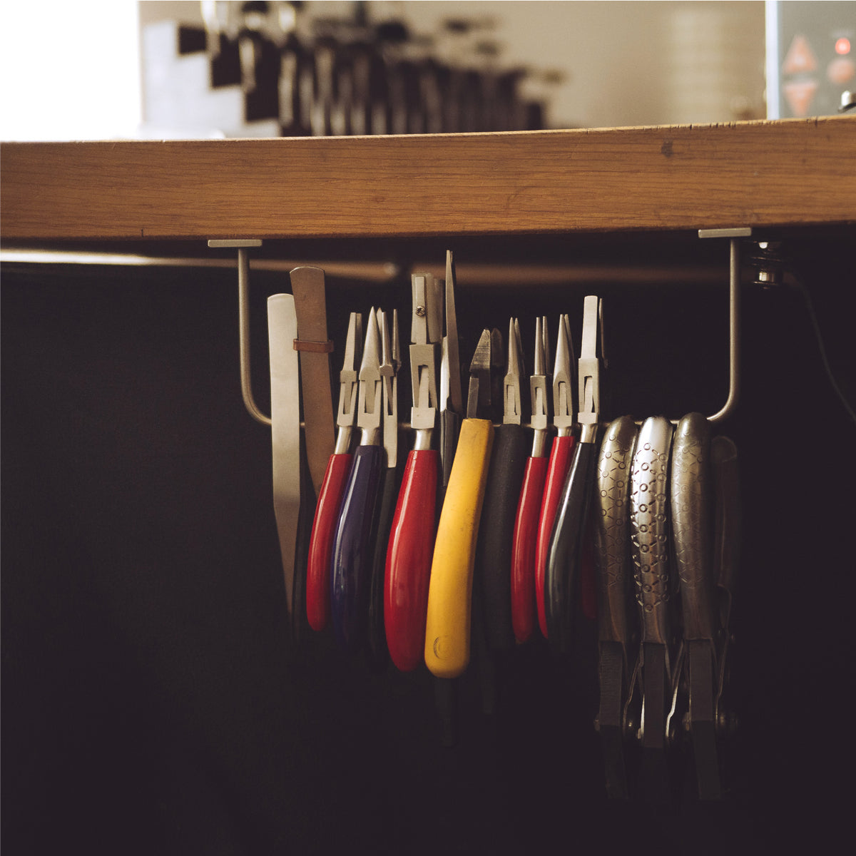 A set of hand tools with colorful handles hanging from a metal rack beneath a wooden surface in a dark workspace.
These tools are used to handmade the objects in italy.