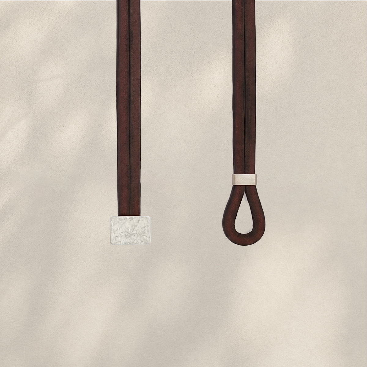 Two dark brown leather straps hanging against a warm beige textured background, one with a square textured silver-toned end and the other with a small silver-toned band near a looped end.