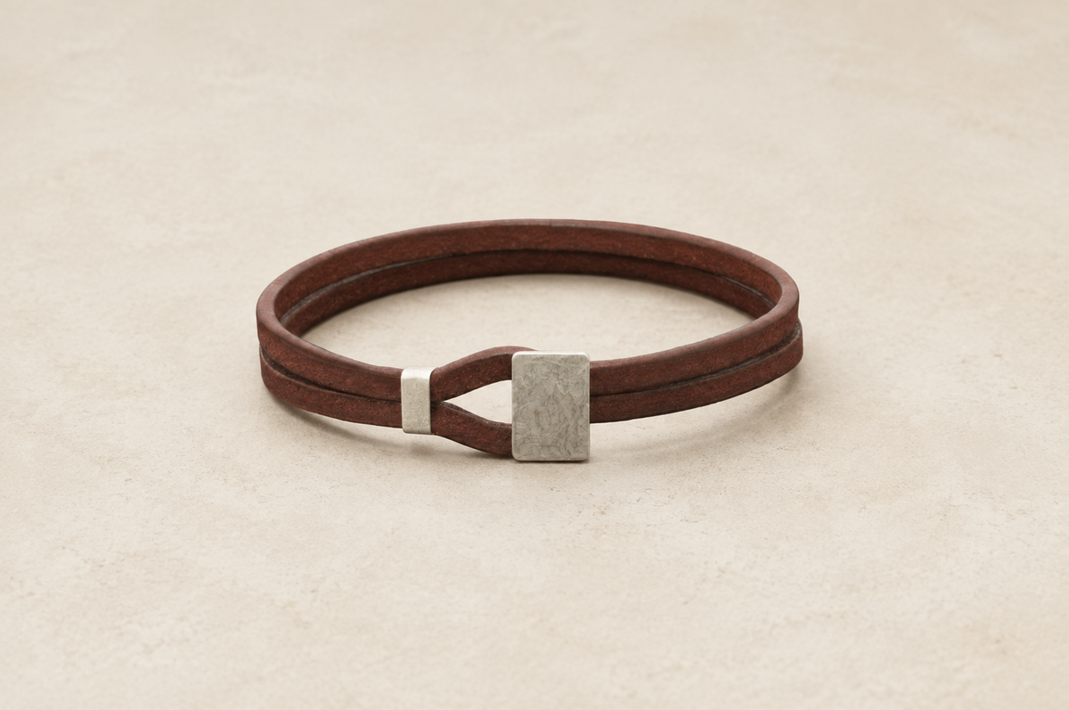 A dark brown leather wrap bracelet with a textured silver metal accent, displayed on a light beige textured background.