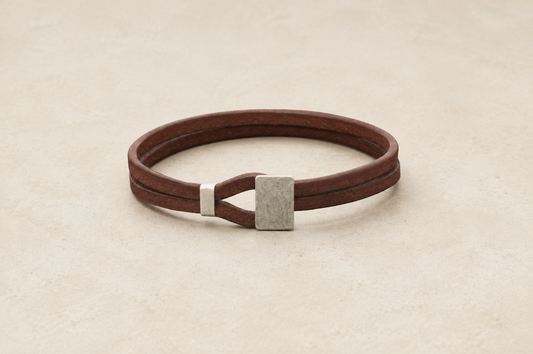 A dark brown leather wrap bracelet with a textured silver metal accent, displayed on a light beige textured background.
