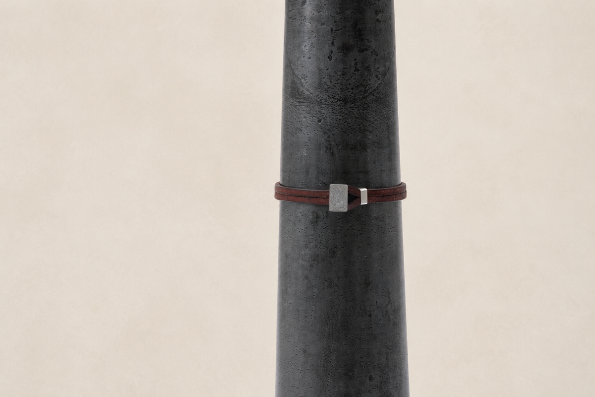 A dark brown leather wrap bracelet with a silver rectangular metal clasp, displayed on a dark gray textured cylindrical stand against a light beige background.