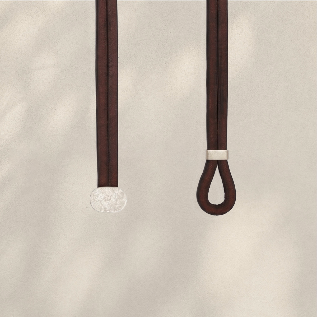 Two dark brown leather strap accessories hanging against a light beige textured background. One strap ends in a round textured silver disc, and the other has a small silver band near the top of a looped end.