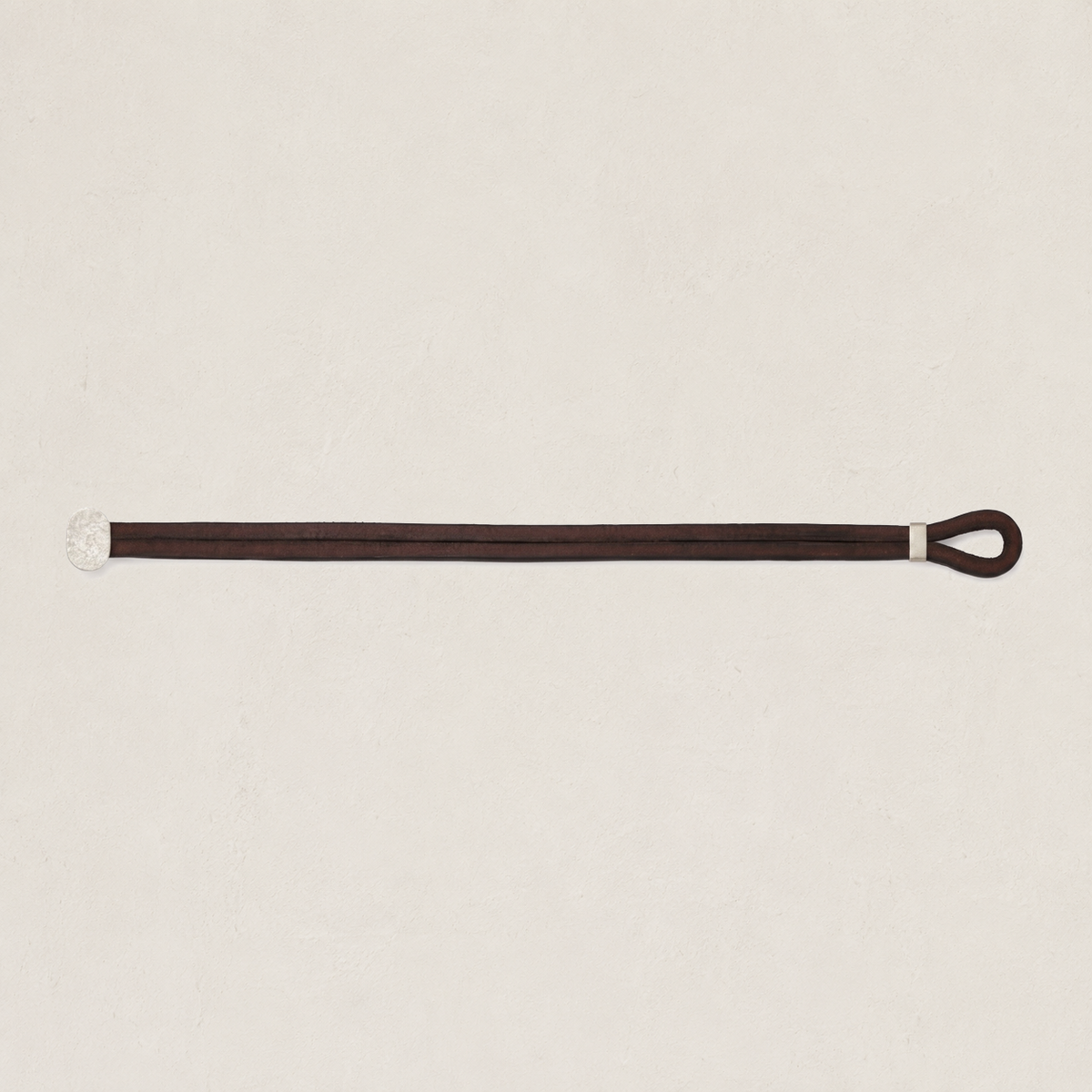 A dark brown leather bracelet with a silver metal clasp and a looped end, laid horizontally on a textured off-white background.