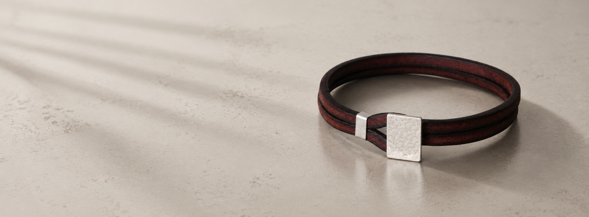 A dark brown leather bangle bracelet with a silver metal connector and square plate, displayed on a light beige textured surface with soft shadows.