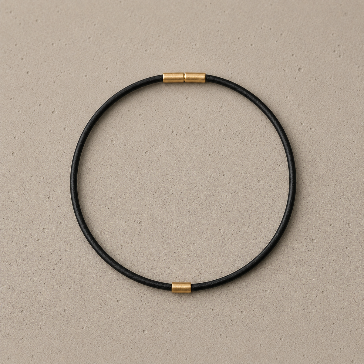 A minimalist black circular bangle anklet with bayonet clasp and 14k solid gold element, displayed on a textured beige background.