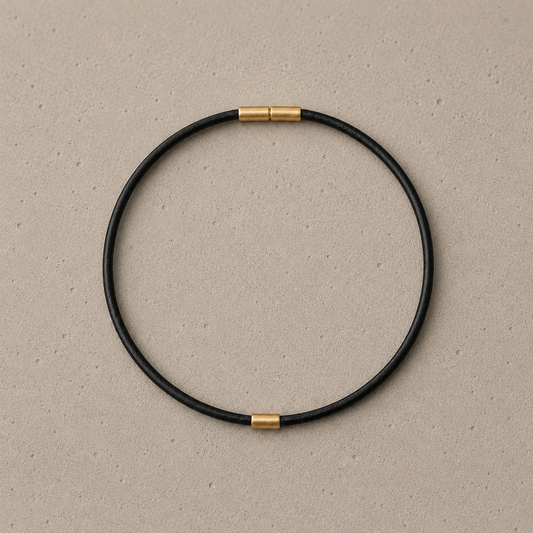 A minimalist black circular bangle anklet with bayonet clasp and 14k solid gold element, displayed on a textured beige background.